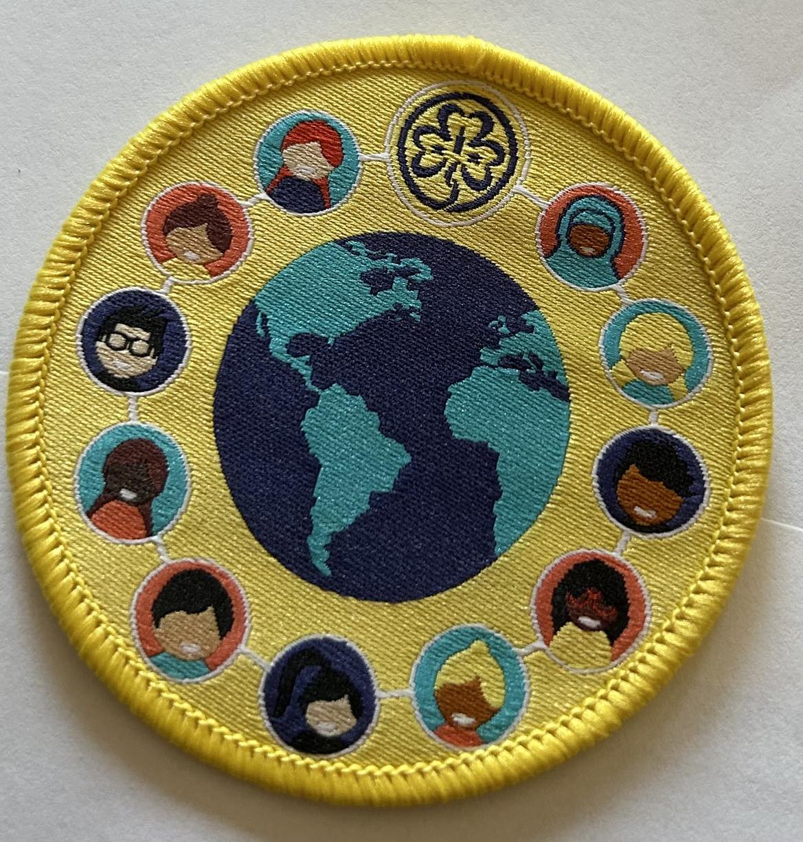 a round badge bound in yellow with part of the world map in the centre with the trefoil and pictures of girls around the outside of the map