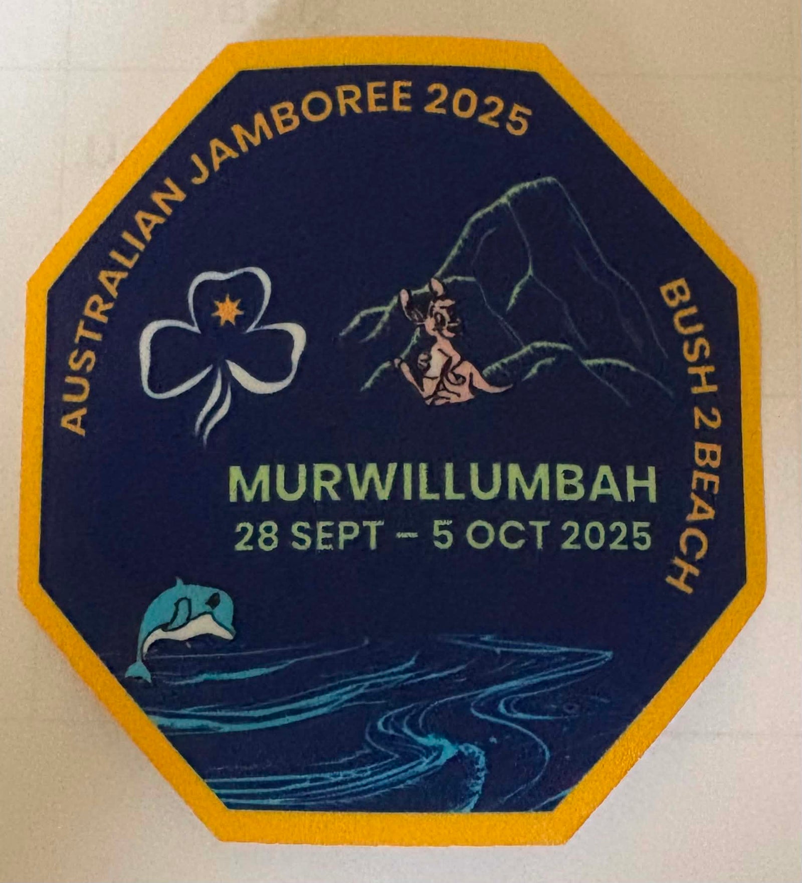 An octagonal commemorative badge with a dark blue background and yellow border, featuring the text 'AUSTRALIAN JAMBOREE 2025' and 'MURWILLUMBAH 28 SEPT - 5 OCT 2025' along with illustrated graphics including a trefoil, mountain, figure, dolphin, and waves.