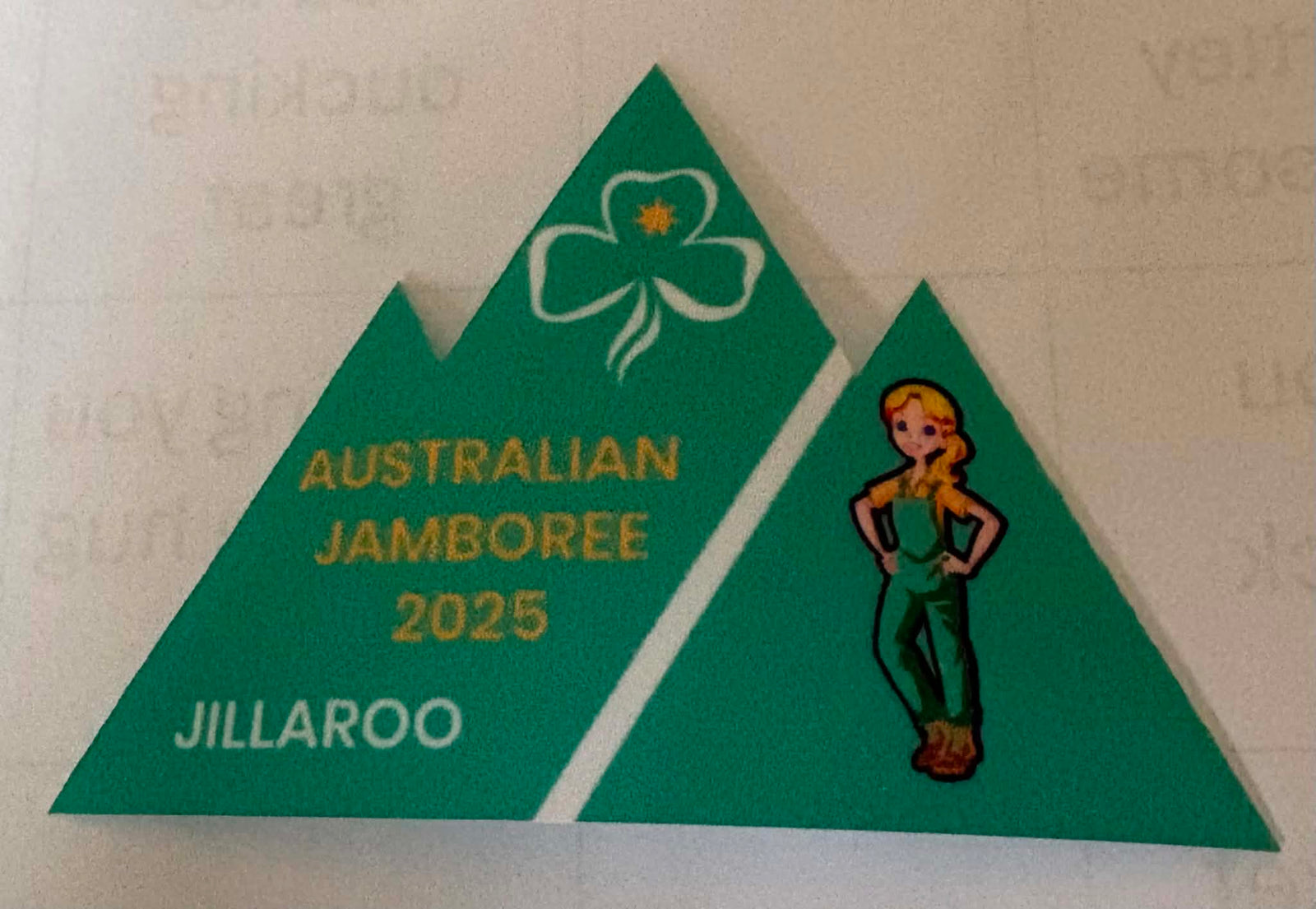 A green triangular commemorative badge for the Australian Jamboree 2025 at Jillaroo, featuring a white trefoil with a gold star, yellow text, and a cartoon woman illustration on a light background.