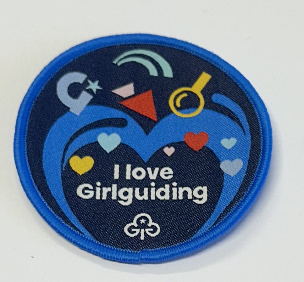 a round blue badge with I love Girlguiding  with hearts and other shapes on it bound in royal blue