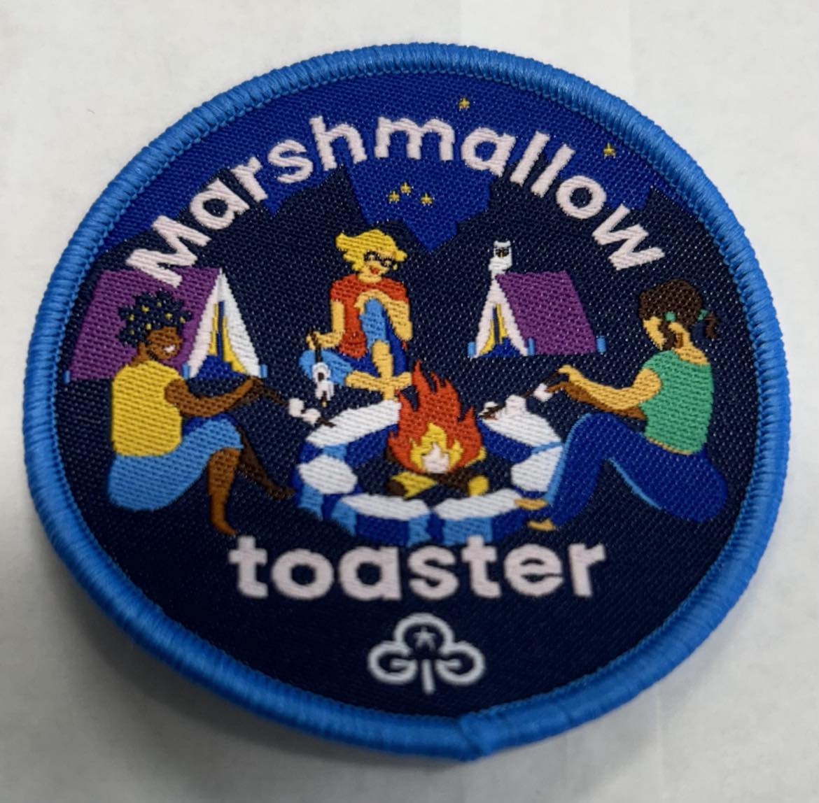 a round badge bound in blue with three girls sitting around a campfire toasting marshmallows with tents in the background