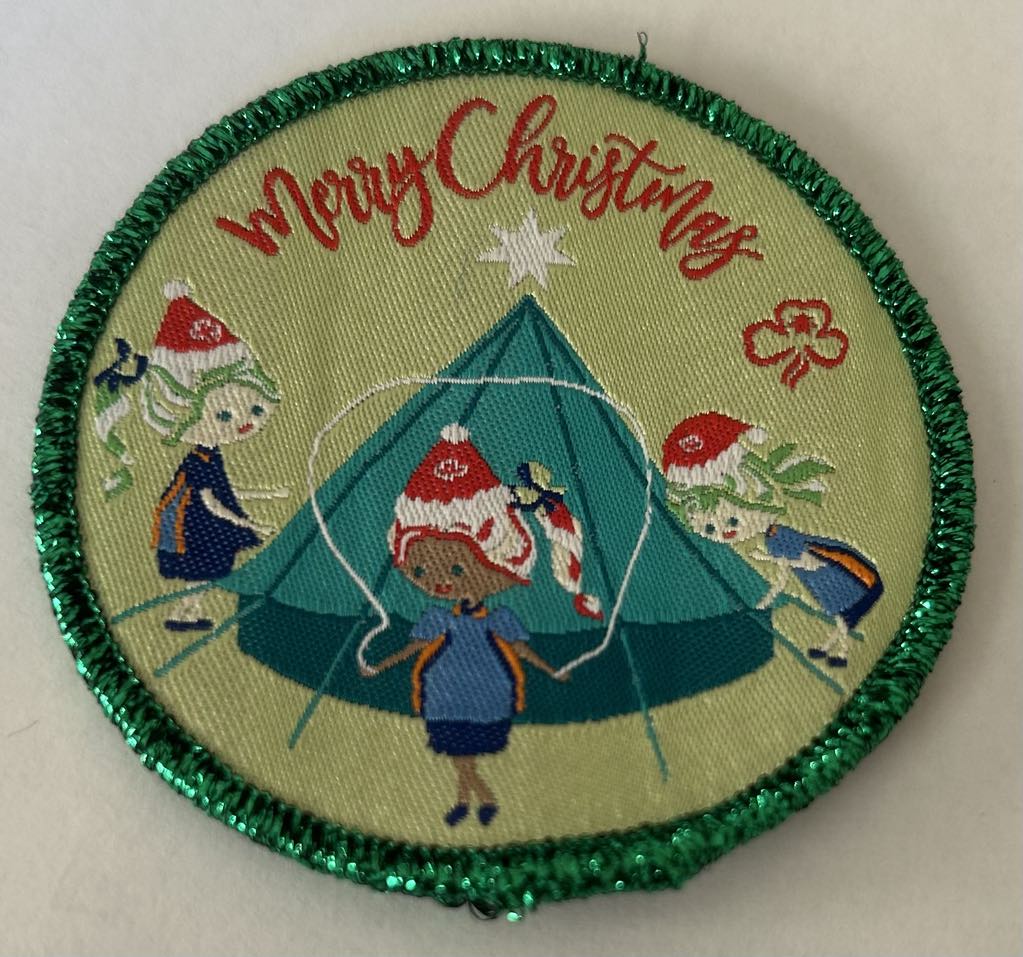 a round green badge bound with dark green metallic binding with the words merry Christmas and three girls with a bell tent