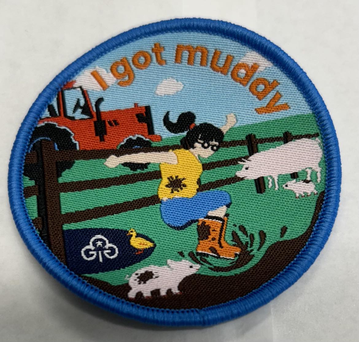 a round badge bound in blue with a tractor, pigs, duck, farm fence and a girl jumping in mud