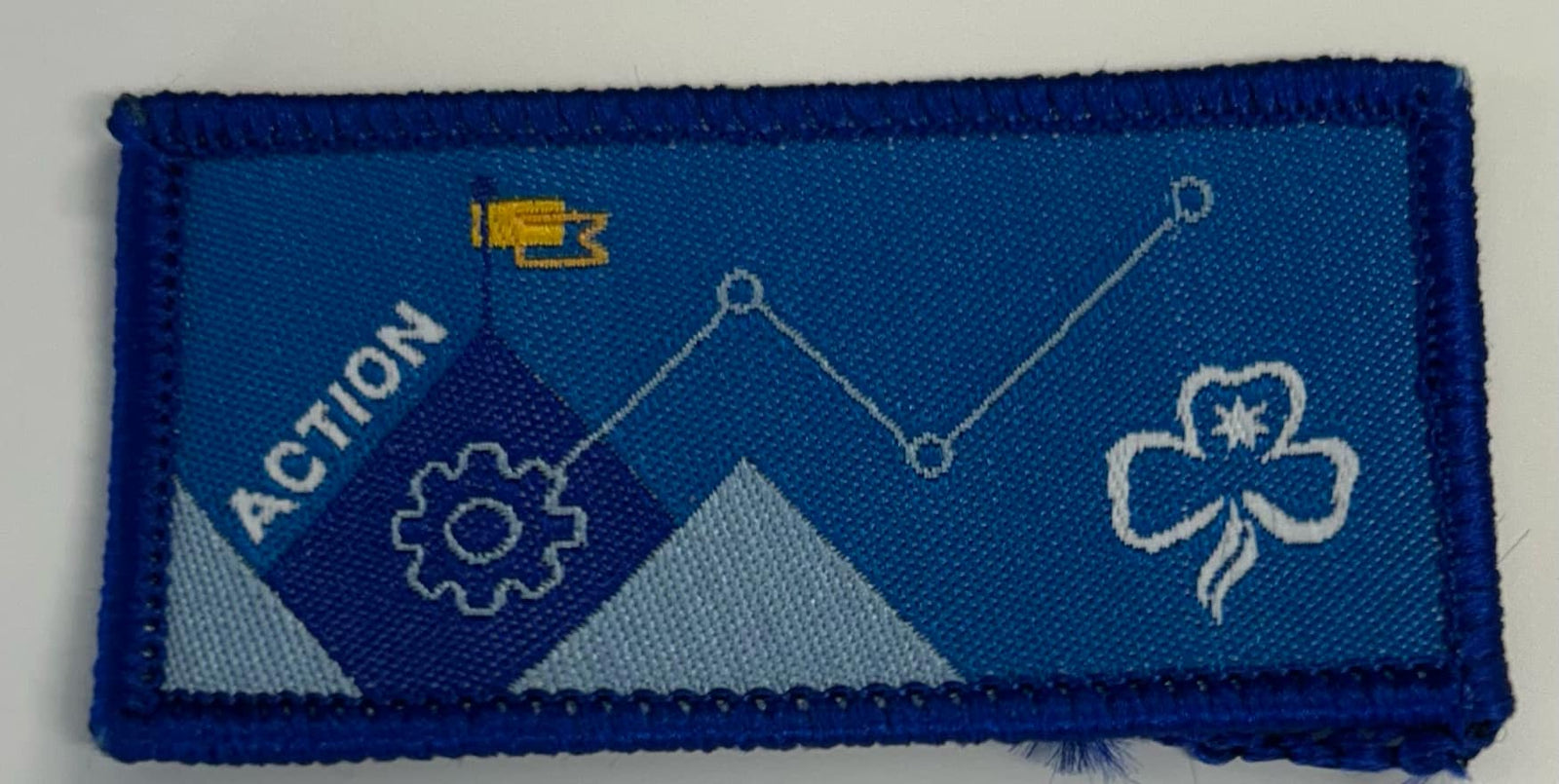 a rectangular bound blue action badge with the new design of a flag, trefoil and flag
