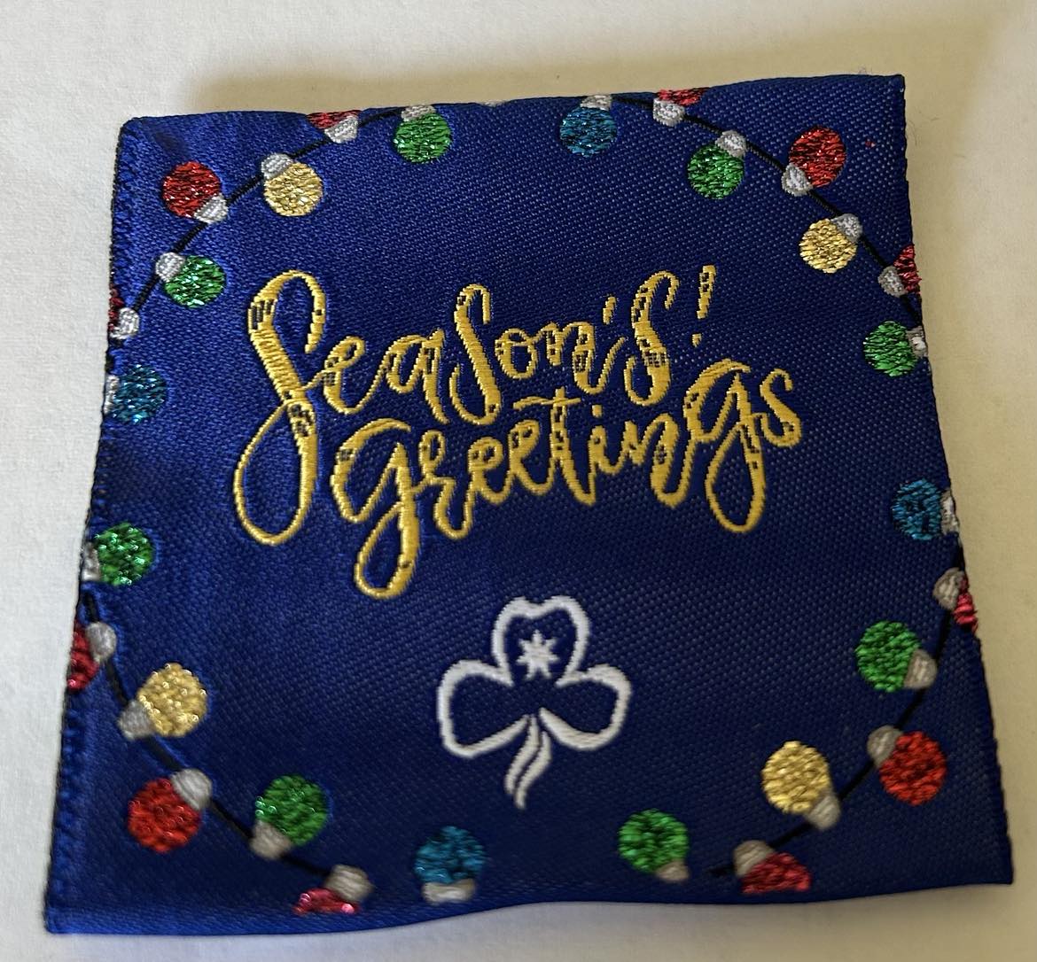 a square unbound badge with a blue background with a circle of Christmas lights with the words seasons greetings in the centre