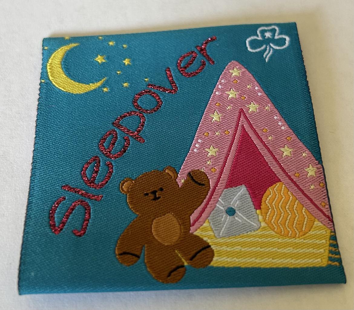 a square blue unbound badge with a teddy bear and a pink tent and the word sleepover in pink and a moon and stars