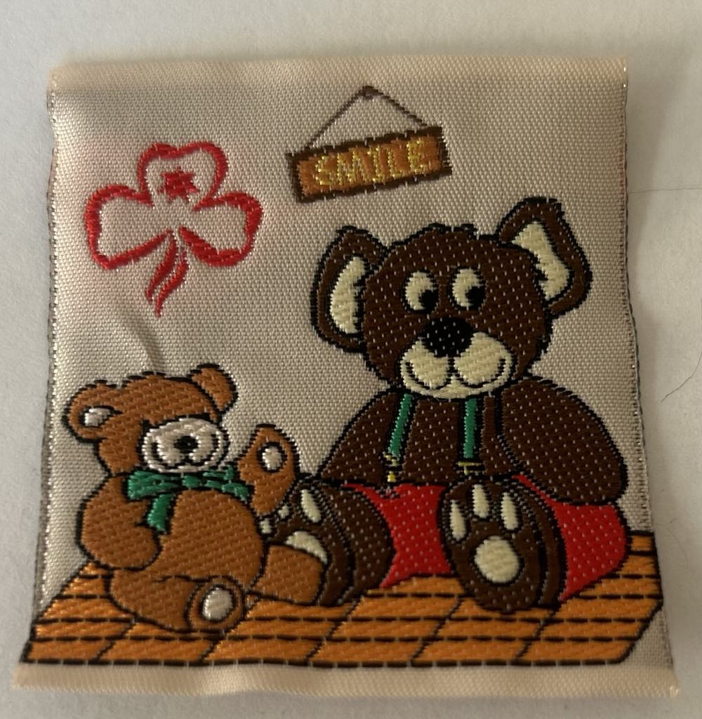 a square unbound badge with two teddy bears sitting on a blanket with the world smile above them