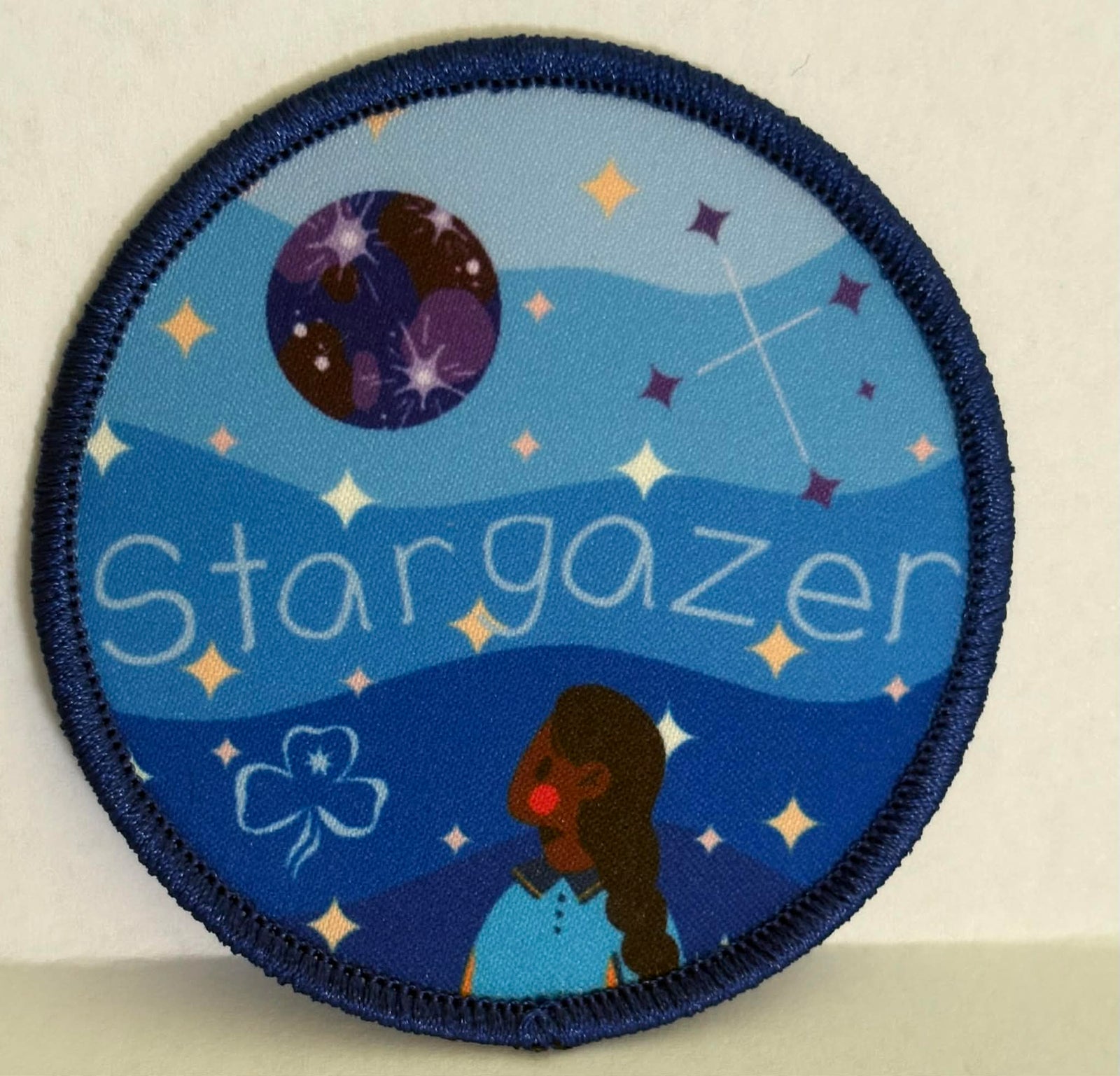 round badge bound in dark blue with 'Stargazer' design featuring a girl looking at a planet and stars.