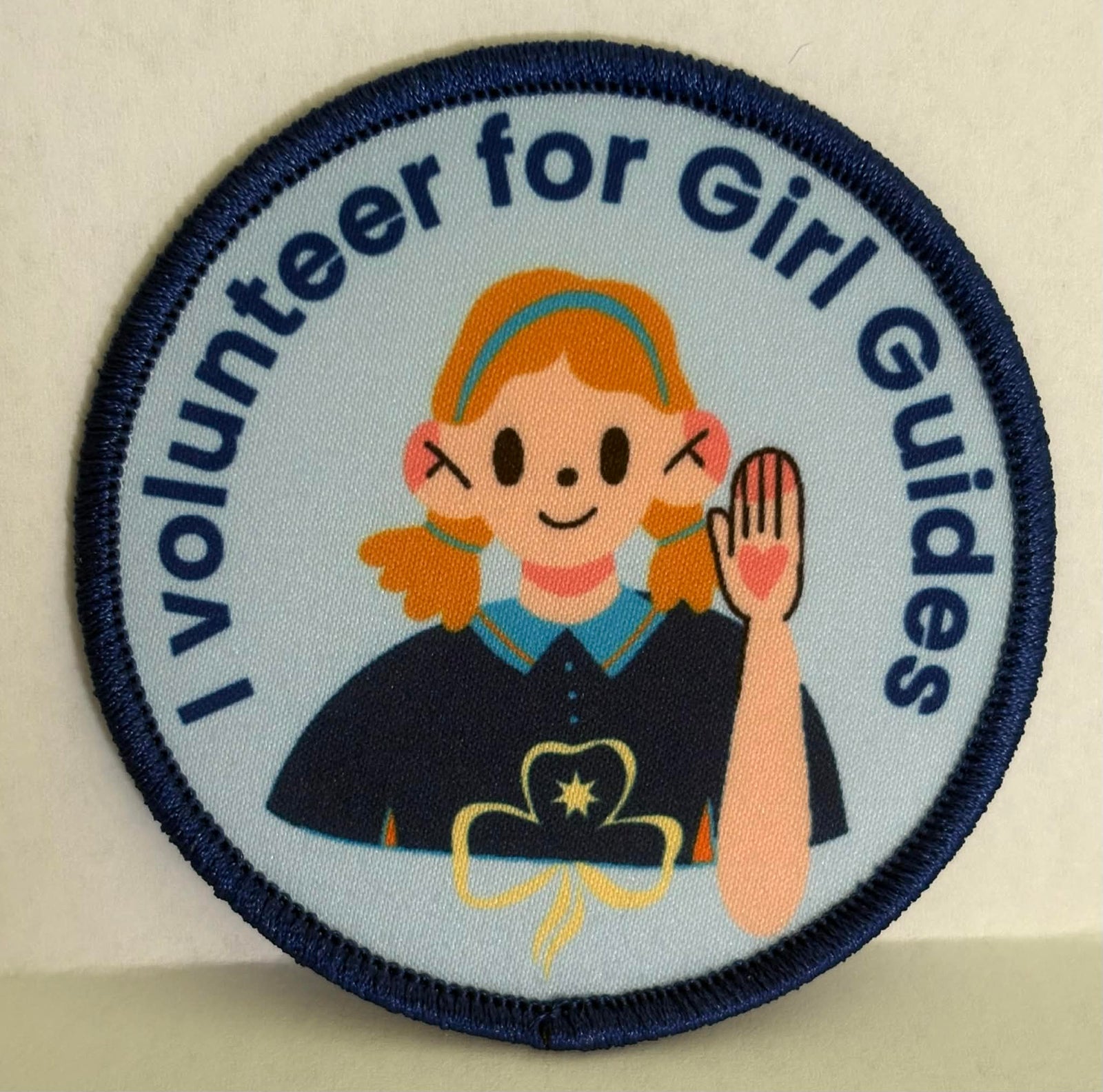 round blue badge bound in dark blue with a female with her hand raised and the words I volunteer for girl guides