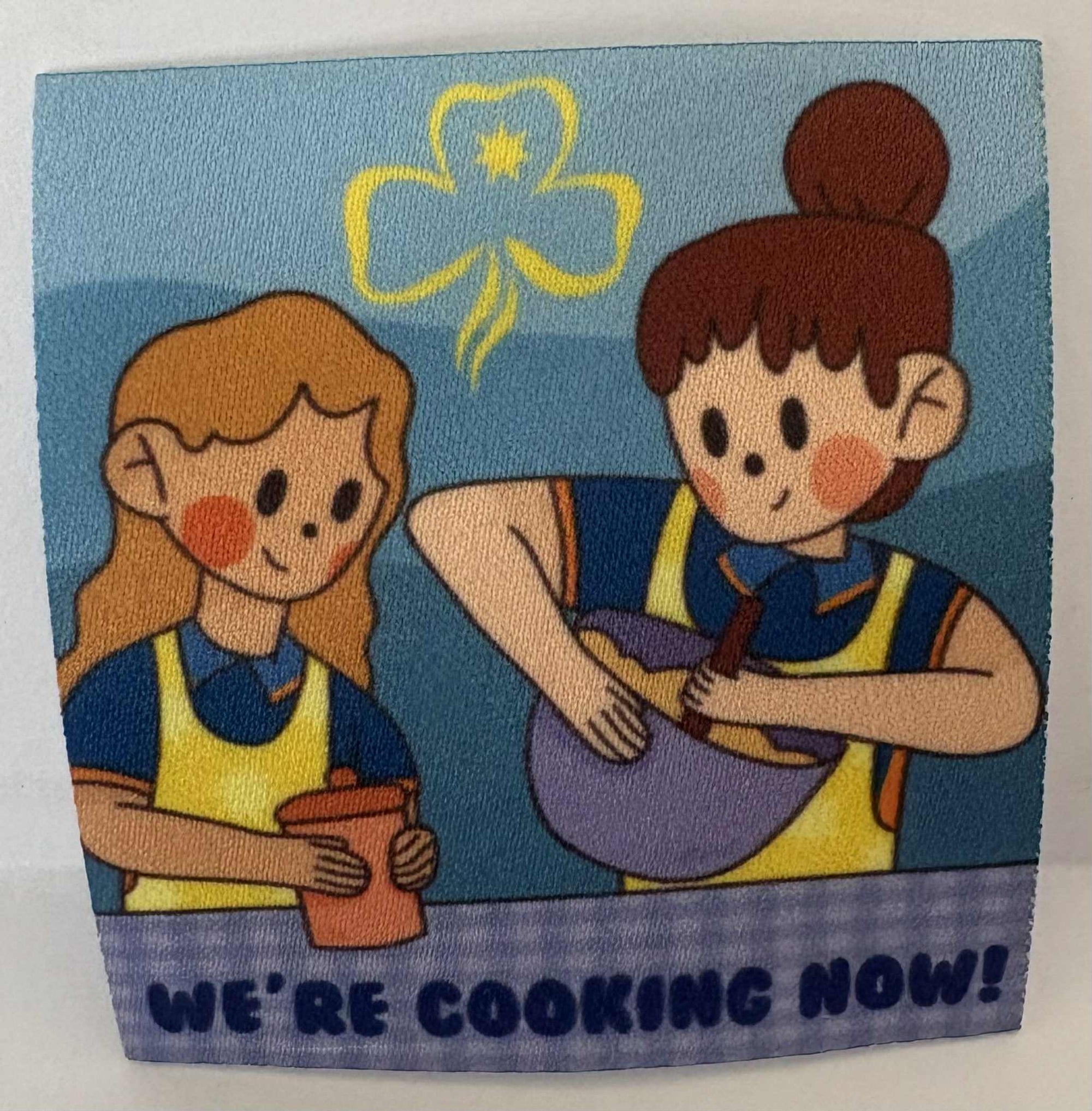 a square unbound badge with two girls with aprons and bowls and a spoon